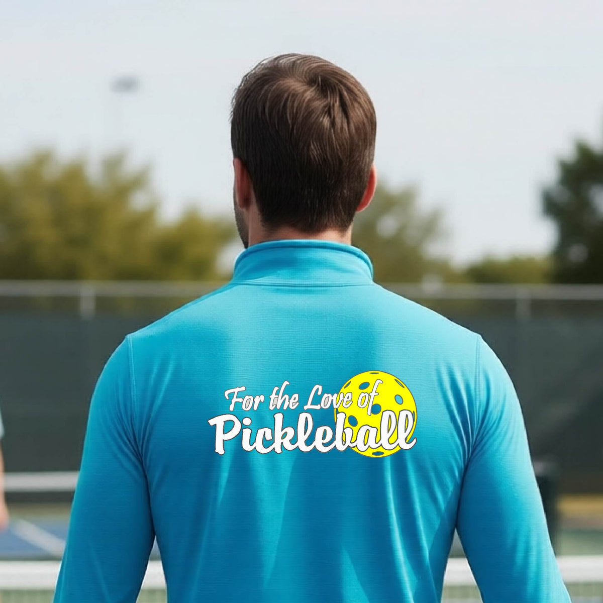 A close-up of a man wearing a bright blue quarter-zip shirt with a pickleball graphic on the left bottom left of ‘FOR THE LOVE OF PICKLEBALL’ in white with a yellow pickleball behind it.  