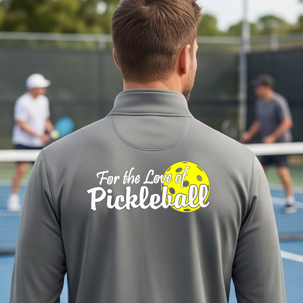 A close-up of a man wearing a gray quarter-zip shirt with a pickleball graphic on the left bottom left of ‘FOR THE LOVE OF PICKLEBALL’ in white with a yellow pickleball behind it.  