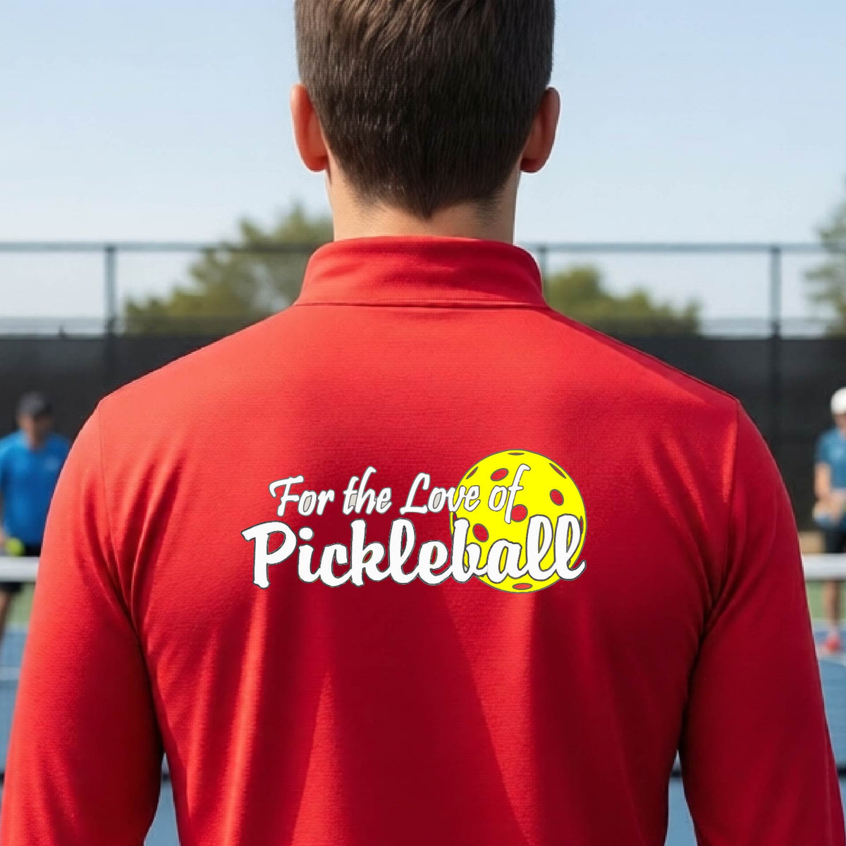 A close-up of a man wearing a red quarter-zip shirt with a pickleball graphic on the left bottom left of ‘FOR THE LOVE OF PICKLEBALL’ in white with a yellow pickleball behind it.  