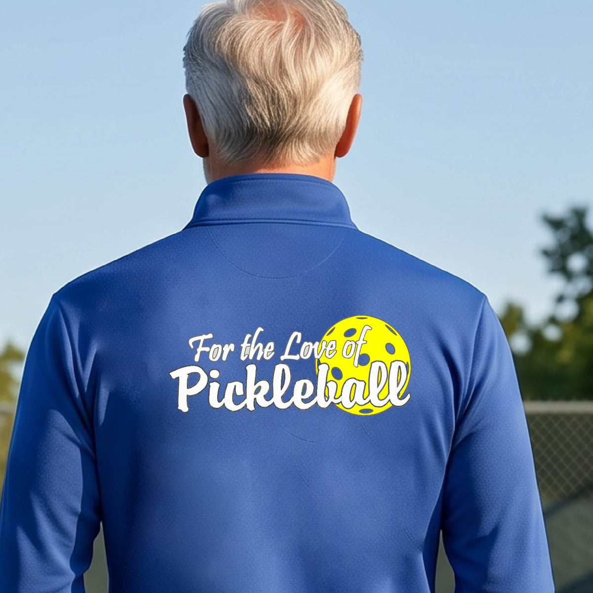 A close-up of a man wearing a royal blue quarter-zip shirt with a pickleball graphic on the left bottom left of ‘FOR THE LOVE OF PICKLEBALL’ in white with a yellow pickleball behind it.  
