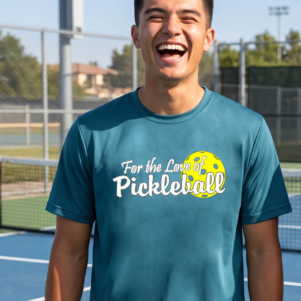 A close-up of a man wearing a teal short-sleeve t-shirt with a pickleball graphic on the left bottom left of ‘FOR THE LOVE OF PICKLEBALL’ in white with a yellow pickleball behind it.  