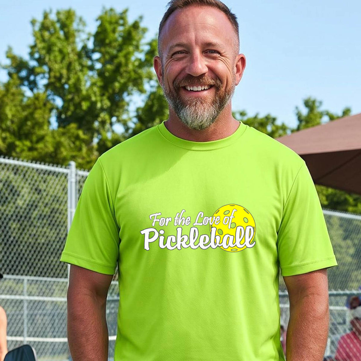 A close-up of a man wearing a lime green short-sleeve t-shirt with a pickleball graphic on the left bottom left of ‘FOR THE LOVE OF PICKLEBALL’ in white with a yellow pickleball behind it.  