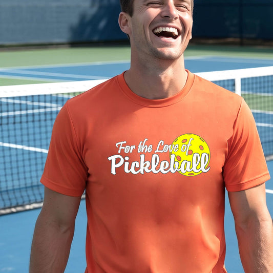 A close-up of a man wearing an orange short-sleeve t-shirt with a pickleball graphic on the left bottom left of ‘FOR THE LOVE OF PICKLEBALL’ in white with a yellow pickleball behind it.  