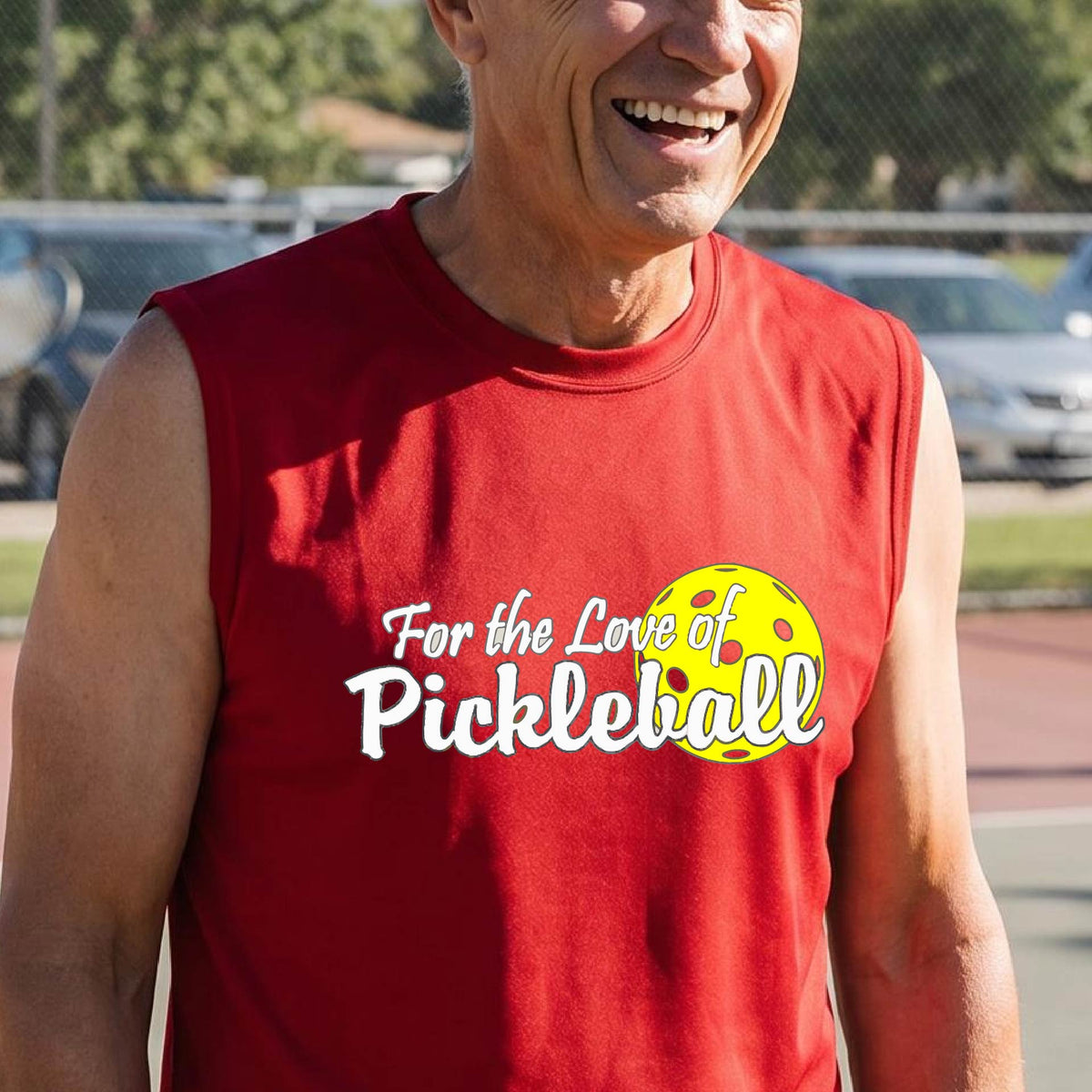 A close-up of a man wearing a red sleeveless t-shirt with a pickleball graphic on the left bottom left of ‘FOR THE LOVE OF PICKLEBALL’ in white with a yellow pickleball behind it.  