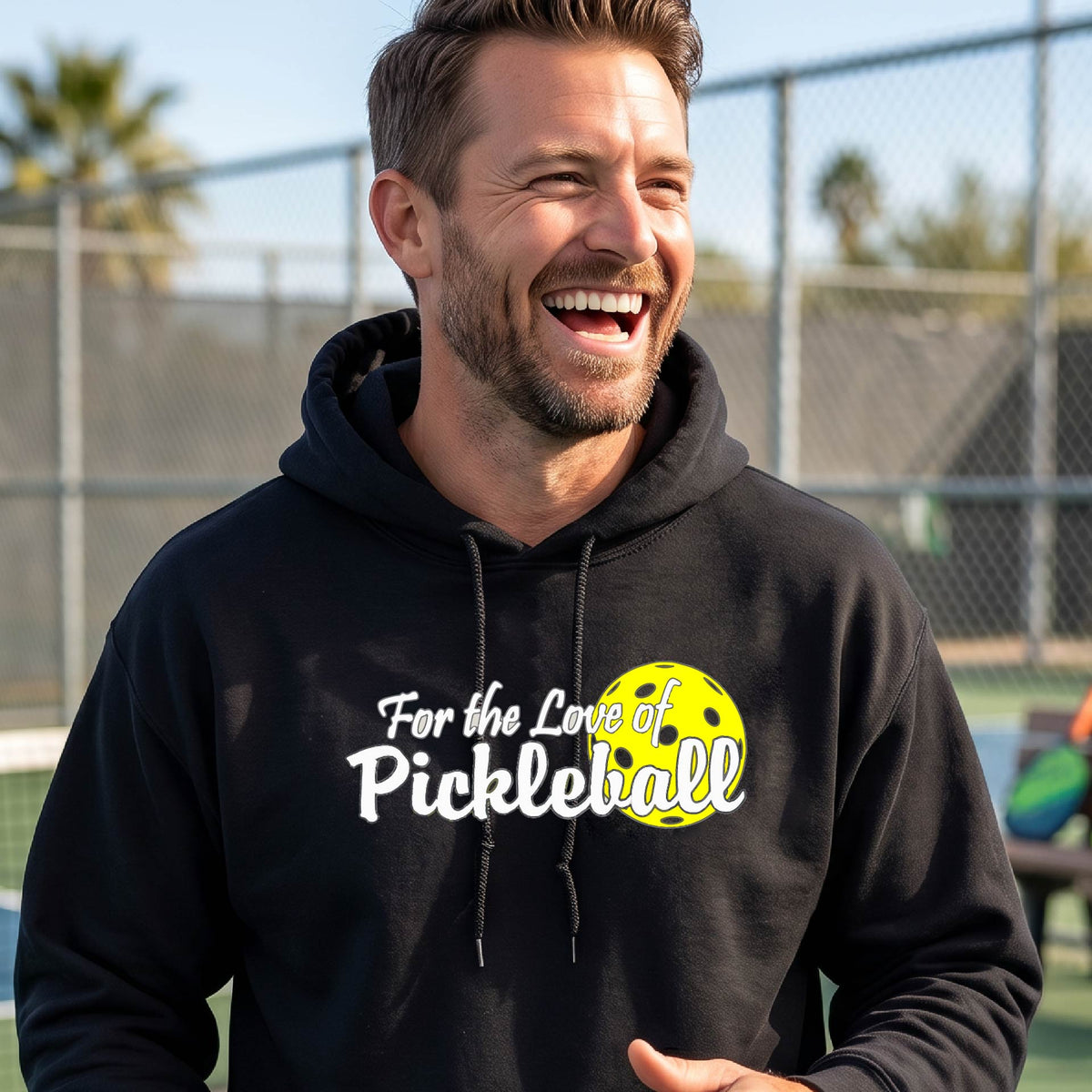 A close-up of a black unisex hoodie pullover with a pickleball graphic of ‘FOR THE LOVE OF PICKLEBALL’ in white with a yellow pickleball behind it graphic on the front.
