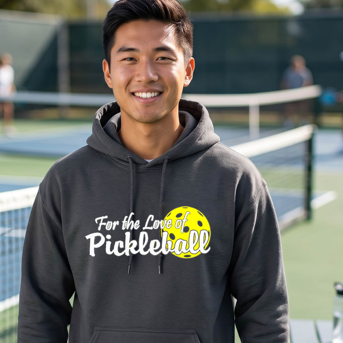 A close-up of a dark gray unisex hoodie pullover with a pickleball graphic of ‘FOR THE LOVE OF PICKLEBALL’ in white with a yellow pickleball behind it graphic on the front.