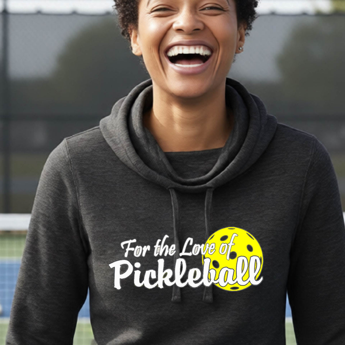 A closeup of a woman wearing a black cowl-neck hoodie pullover with a pickleball graphic on the left bottom left of ‘FOR THE LOVE OF PICKLEBALL’ in white with a yellow pickleball behind it.  