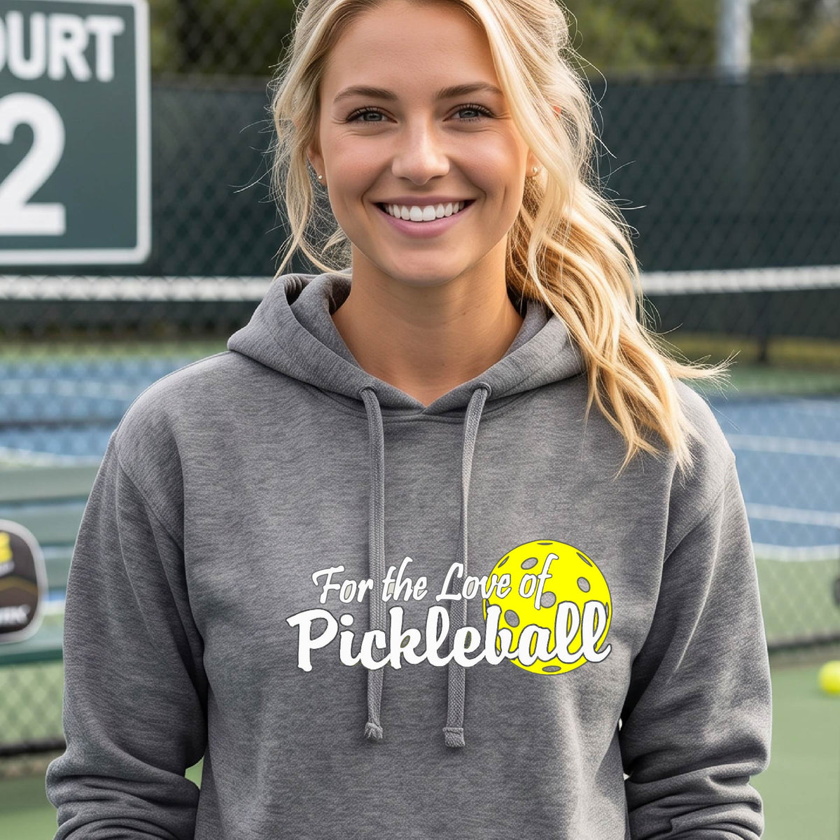 A closeup of a woman wearing a gray hoodie pullover with a pickleball graphic on the left bottom left of ‘FOR THE LOVE OF PICKLEBLL’ in white with a yellow pickleball behind it.  