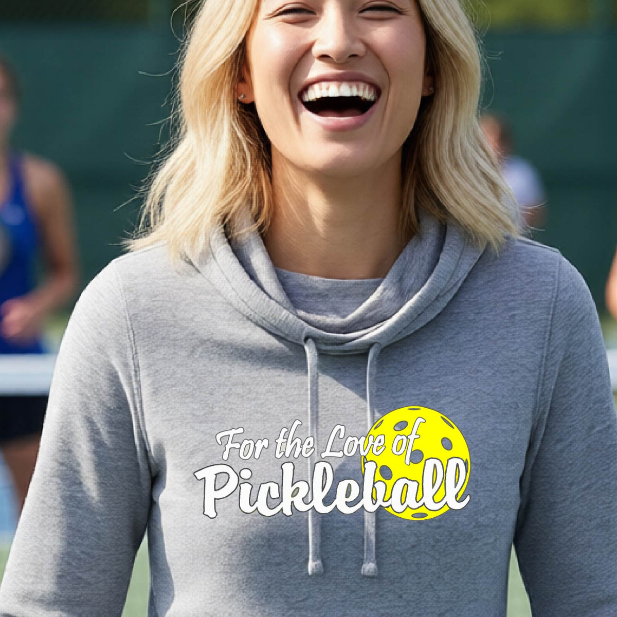 A closeup of a woman wearing a gray cowl-neck hoodie pullover with a pickleball graphic on the left bottom left of ‘FOR THE LOVE OF PICKLEBALL’ in white with a yellow pickleball behind it.  
