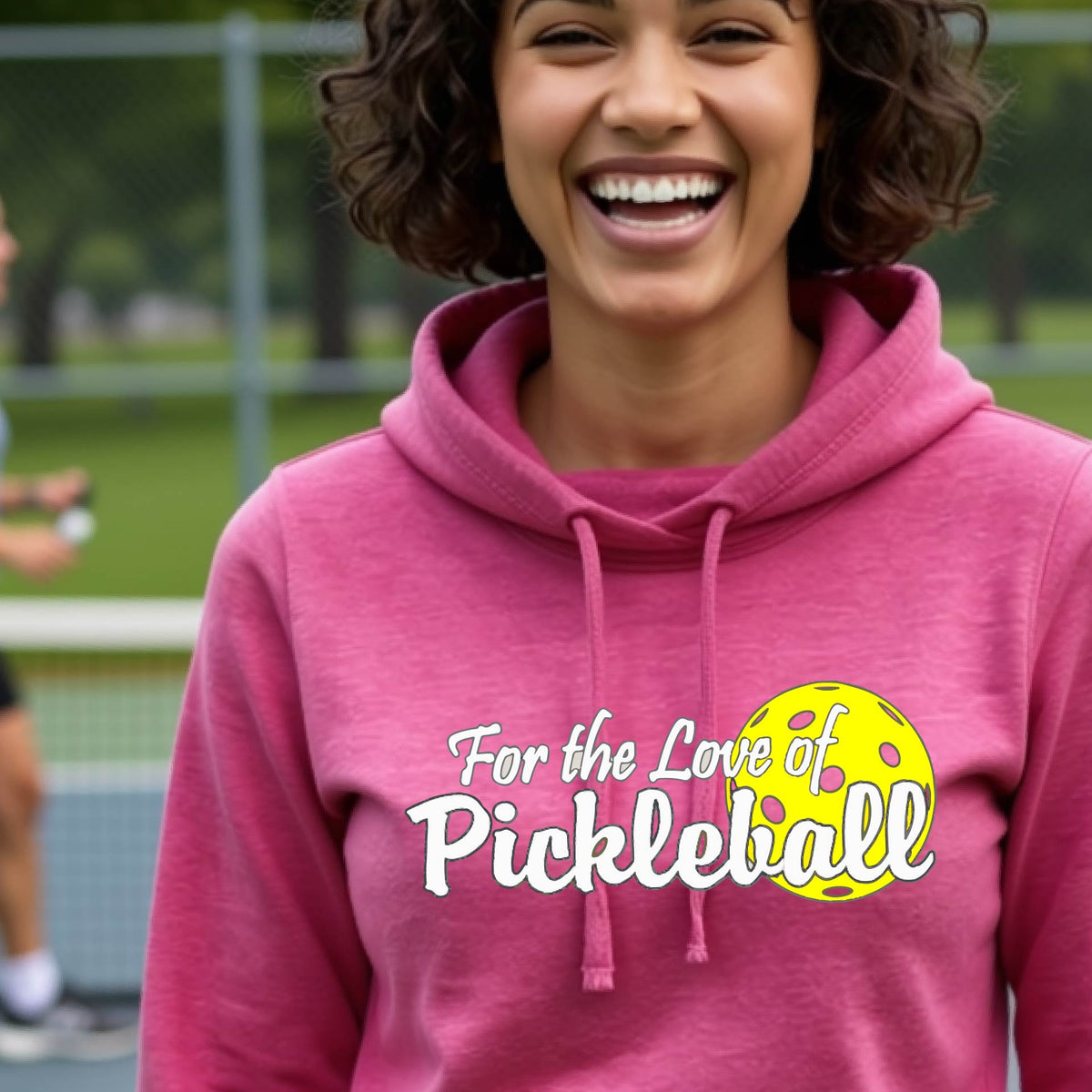 A closeup of a woman wearing a pink cowl-neck hoodie pullover with a pickleball graphic on the left bottom left of ‘FOR THE LOVE OF PICKLEBALL’ in white with a yellow pickleball behind it.  
