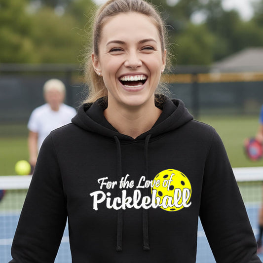 A closeup of a woman wearing a black hoodie pullover with a pickleball graphic on the left bottom left of ‘FOR THE LOVE OF PICKLEBLL’ in white with a yellow pickleball behind it.  