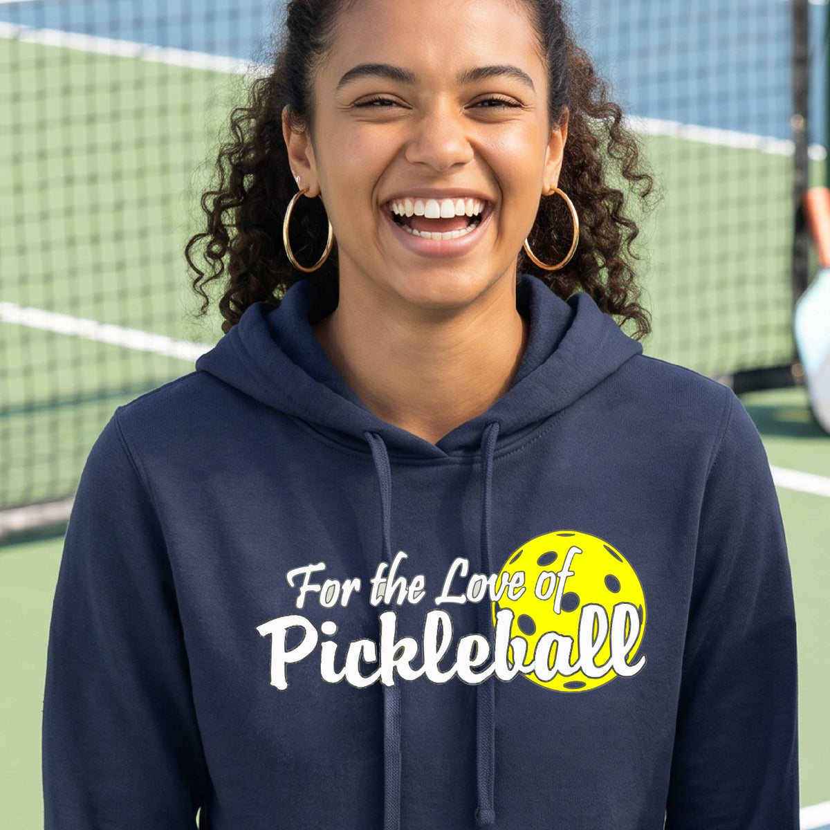 A closeup of a woman wearing a navy blue hoodie pullover with a pickleball graphic on the left bottom left of ‘FOR THE LOVE OF PICKLEBLL’ in white with a yellow pickleball behind it.  