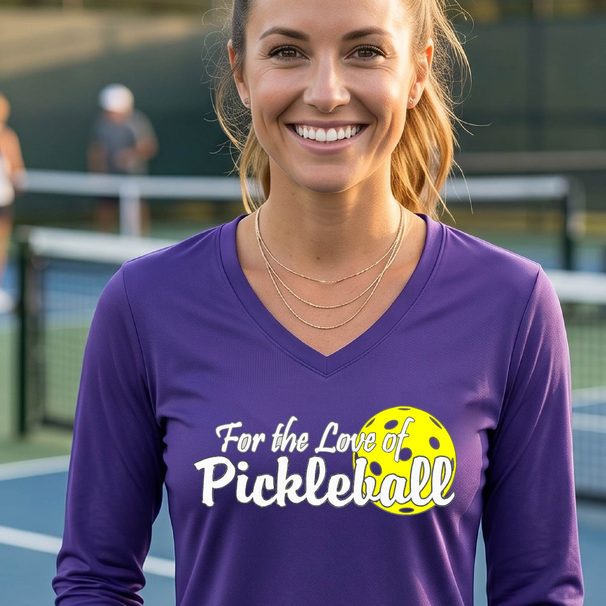 A close-up of a womens purple long-sleeve v-neck t-shirt with a pickleball graphic of ‘FOR THE LOVE OF PICKLEBALL’ in white with a yellow pickleball behind it graphic on the front.