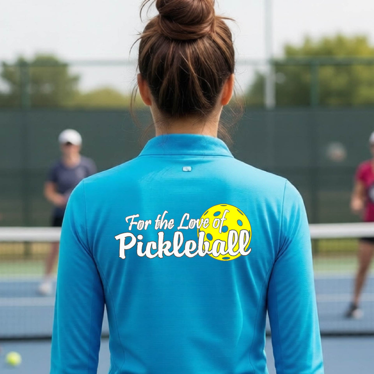 A closeup of a woman wearing a bright blue quarter-zip shirt with a pickleball graphic on the left bottom left of ‘FOR THE LOVE OF PICKLEBLL’ in white with a yellow pickleball behind it.  
