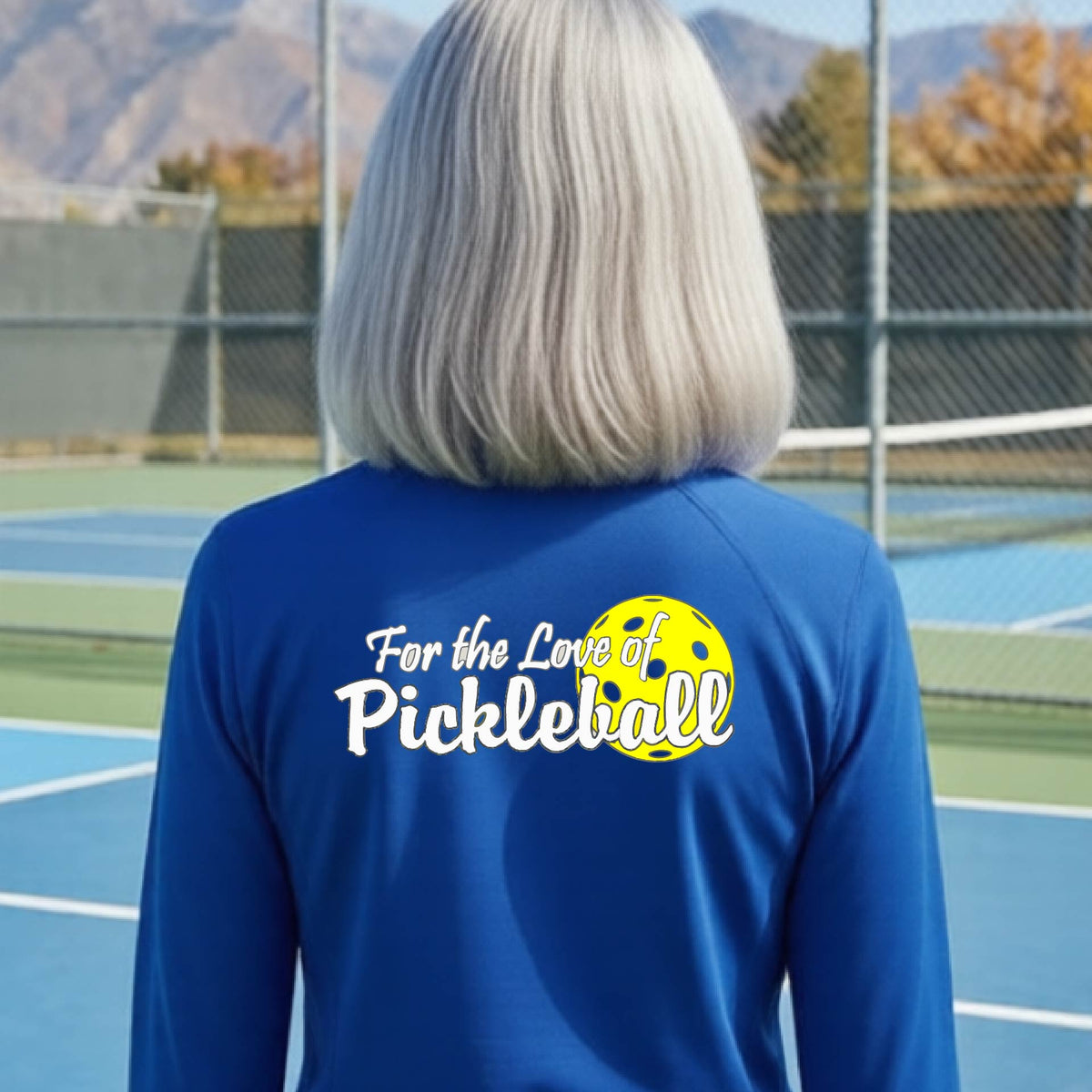 A closeup of a woman wearing a royal blue quarter-zip shirt with a pickleball graphic on the left bottom left of ‘FOR THE LOVE OF PICKLEBLL’ in white with a yellow pickleball behind it.  
