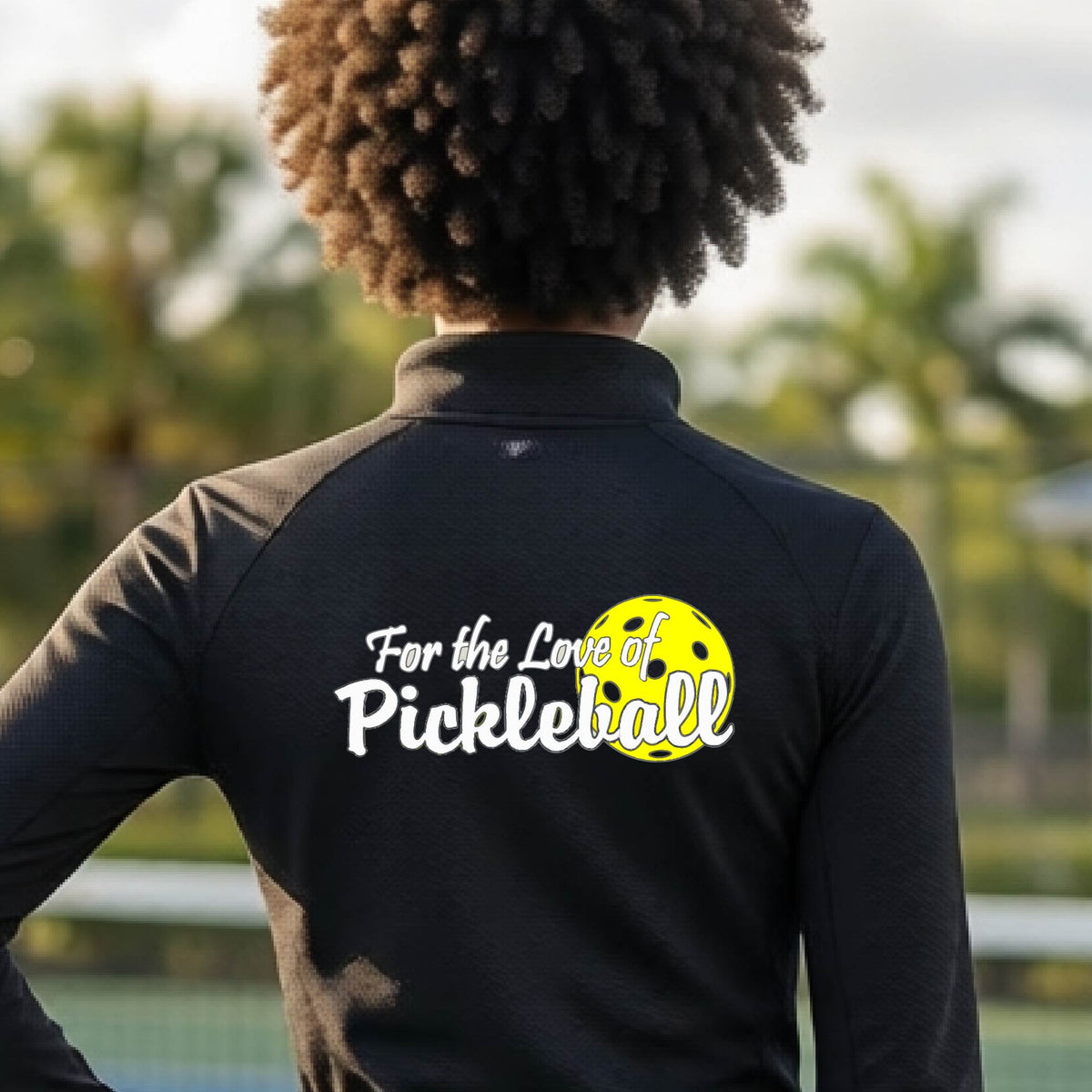 A closeup of a woman wearing a black quarter-zip shirt with a pickleball graphic on the left bottom left of ‘FOR THE LOVE OF PICKLEBLL’ in white with a yellow pickleball behind it.  
