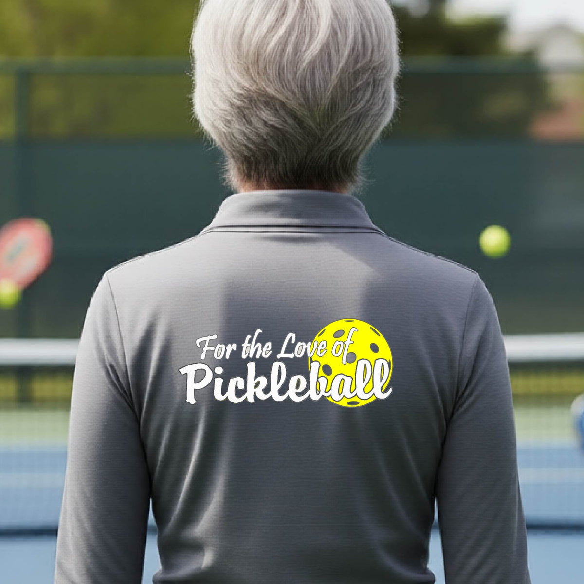 A closeup of a woman wearing a gray quarter-zip shirt with a pickleball graphic on the left bottom left of ‘FOR THE LOVE OF PICKLEBLL’ in white with a yellow pickleball behind it.  
