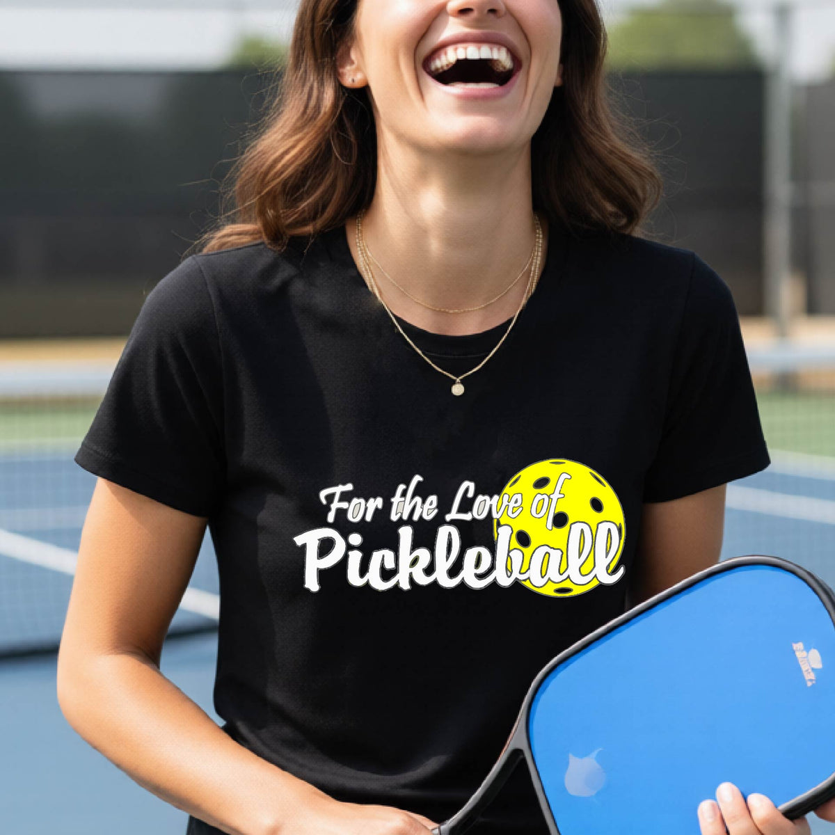 A closeup of a woman wearing a black short-sleeve athletic shirt with a pickleball graphic on the left bottom left of ‘FOR THE LOVE OF PICKLEBLL’ in white with a yellow pickleball behind it.  