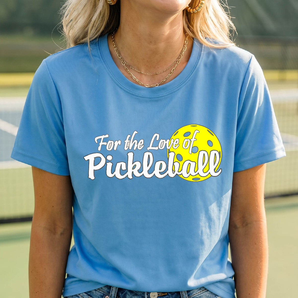 A closeup of a woman wearing a blue short-sleeve athletic shirt with a pickleball graphic on the left bottom left of ‘FOR THE LOVE OF PICKLEBLL’ in white with a yellow pickleball behind it.  