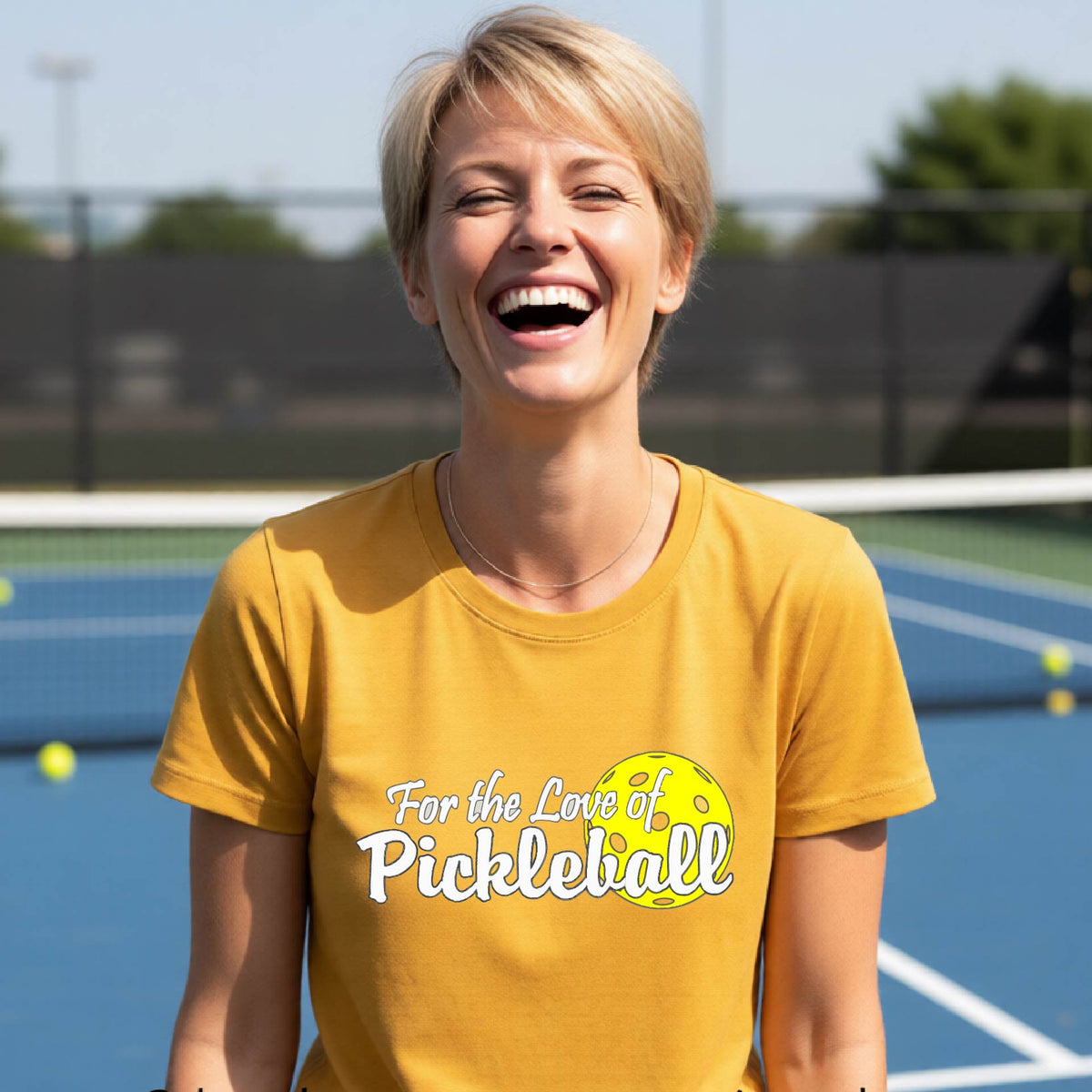 A closeup of a woman wearing a gold short-sleeve athletic shirt with a pickleball graphic on the left bottom left of ‘FOR THE LOVE OF PICKLEBLL’ in white with a yellow pickleball behind it.  