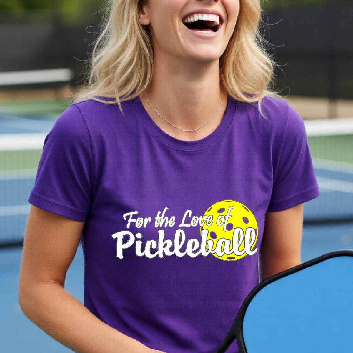A closeup of a woman wearing a purple short-sleeve athletic shirt with a pickleball graphic on the left bottom left of ‘FOR THE LOVE OF PICKLEBLL’ in white with a yellow pickleball behind it.  