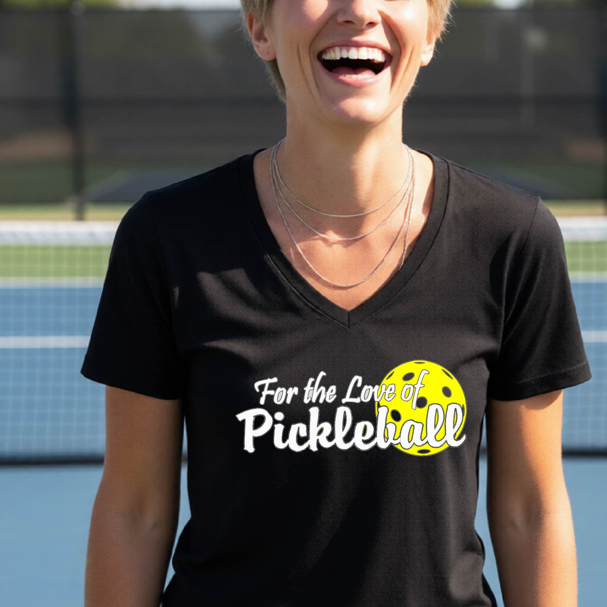 A close-up of a womens black short-sleeve v-neck t-shirt with a pickleball graphic of ‘FOR THE LOVE OF PICKLEBALL’ in white with a yellow pickleball behind it graphic on the front.
