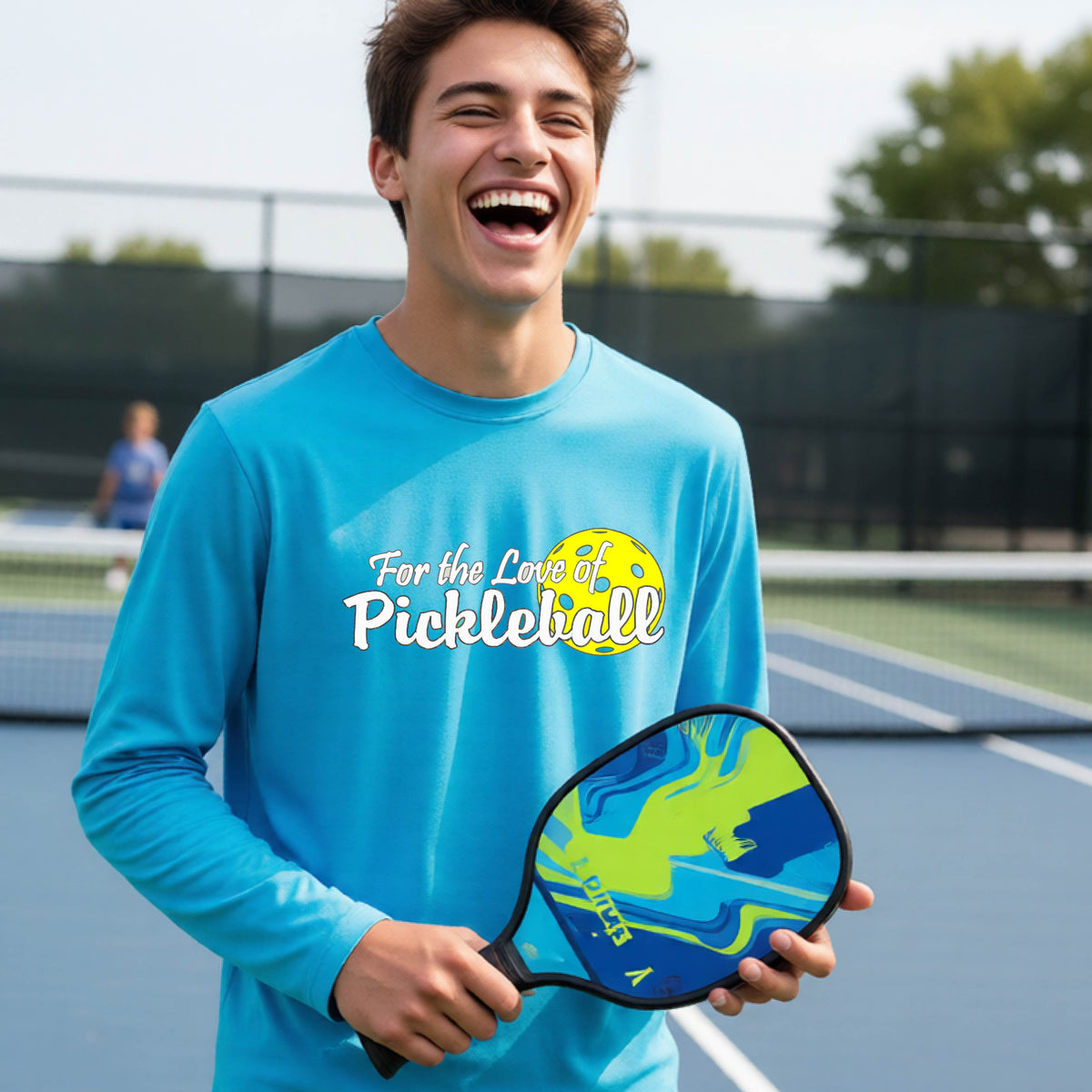 A closeup of a young person wearing a bright blue long-sleeve athletic shirt with a pickleball graphic on the left bottom left of ‘FOR THE LOVE OF PICKLEBLL’ in white with a yellow pickleball behind it.  