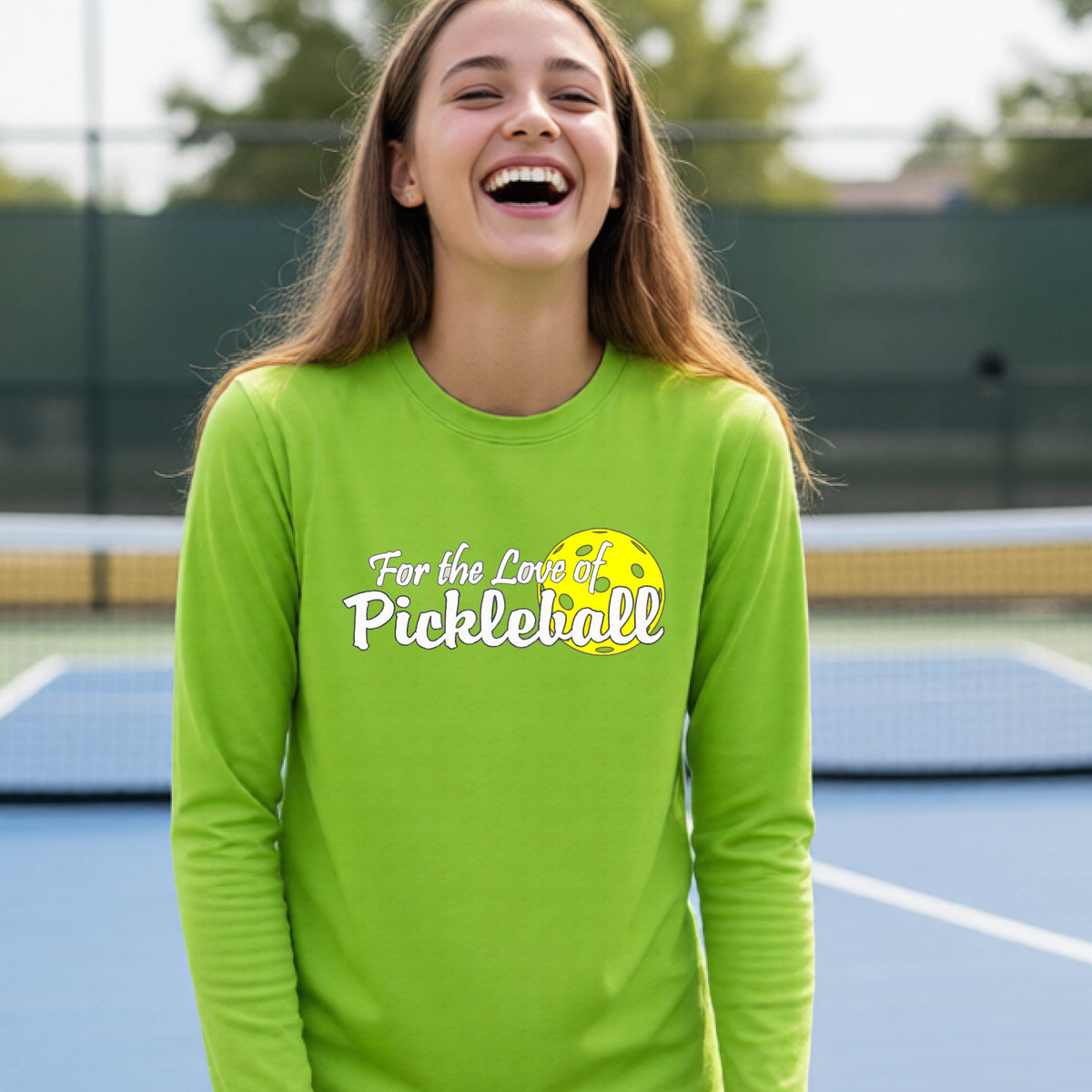 A closeup of a young person wearing a lime green long-sleeve athletic shirt with a pickleball graphic on the left bottom left of ‘FOR THE LOVE OF PICKLEBLL’ in white with a yellow pickleball behind it.  