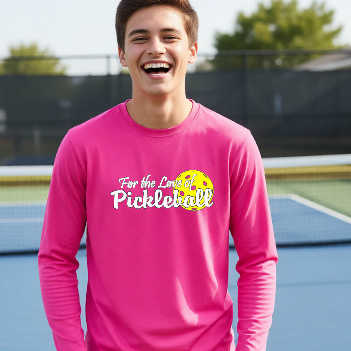 A closeup of a young person wearing a pink long-sleeve athletic shirt with a pickleball graphic on the left bottom left of ‘FOR THE LOVE OF PICKLEBLL’ in white with a yellow pickleball behind it.  