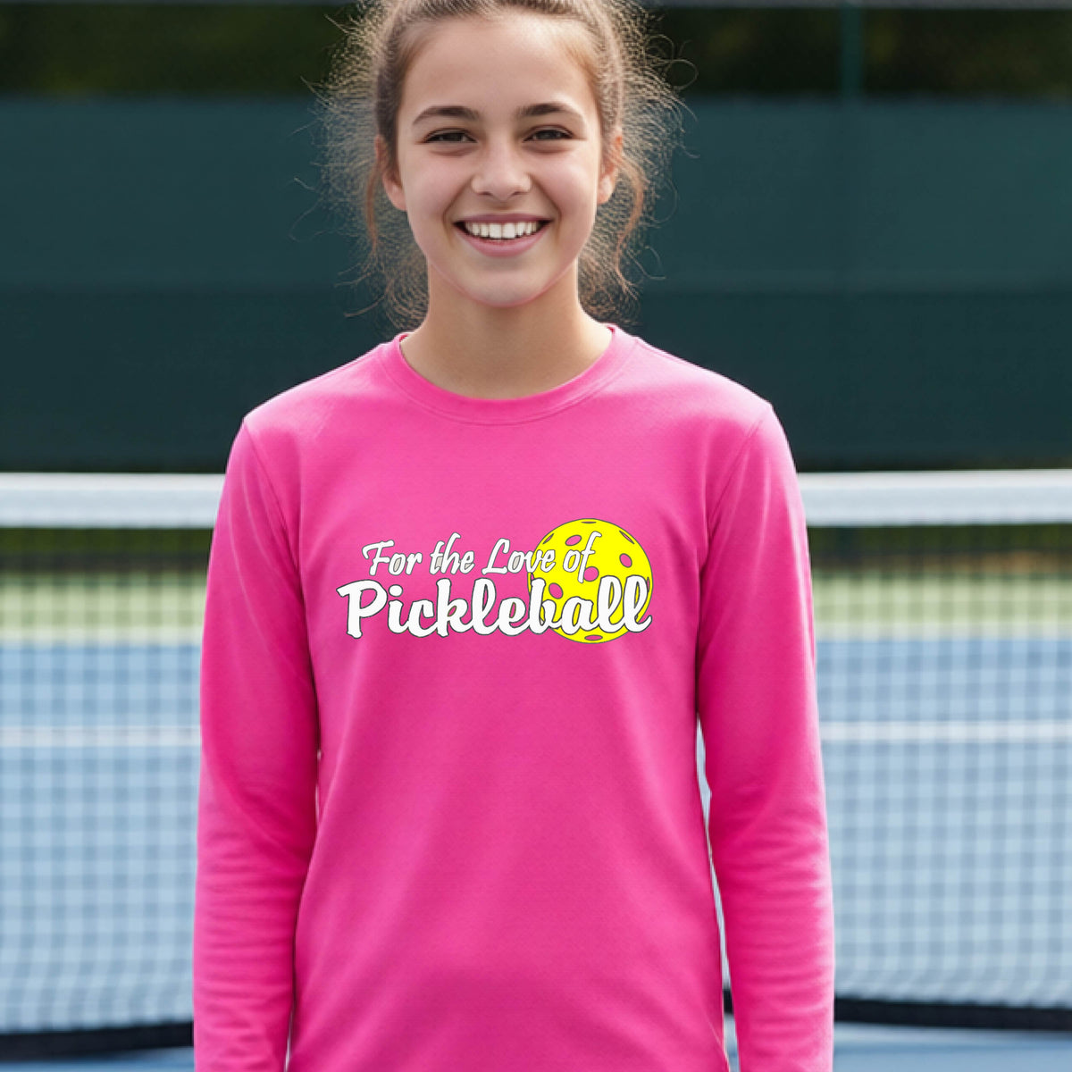 A closeup of a young person wearing a pink long-sleeve athletic shirt with a pickleball graphic on the left bottom left of ‘FOR THE LOVE OF PICKLEBLL’ in white with a yellow pickleball behind it.  