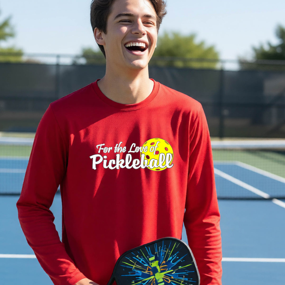 A closeup of a young person wearing a red long-sleeve athletic shirt with a pickleball graphic on the left bottom left of ‘FOR THE LOVE OF PICKLEBLL’ in white with a yellow pickleball behind it.  