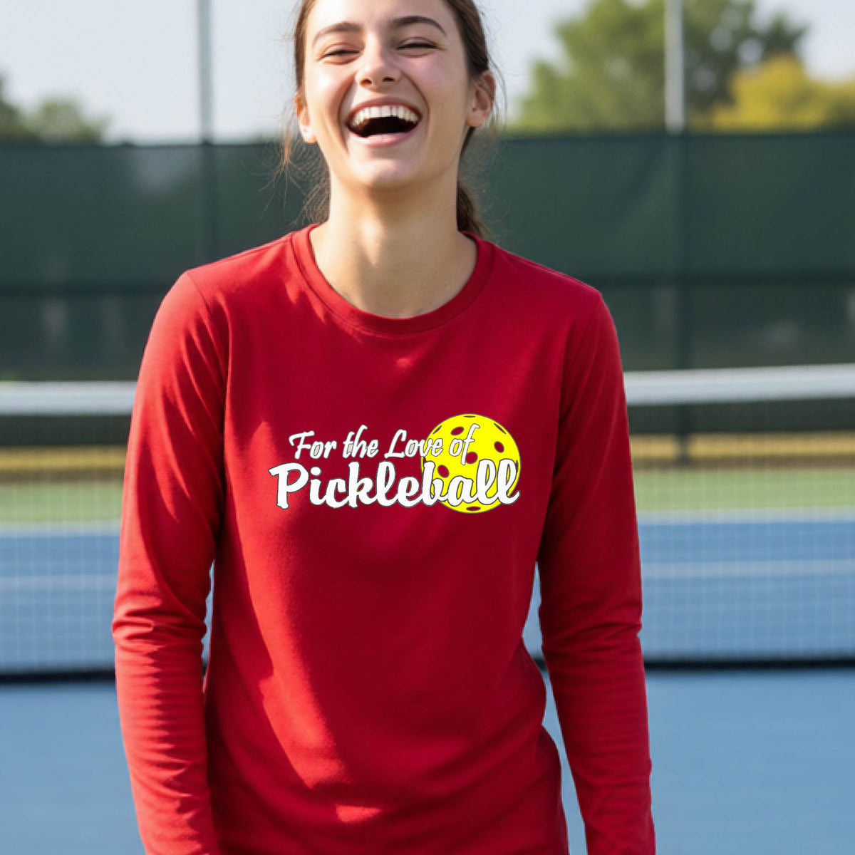 A closeup of a young person wearing a red long-sleeve athletic shirt with a pickleball graphic on the left bottom left of ‘FOR THE LOVE OF PICKLEBLL’ in white with a yellow pickleball behind it.  