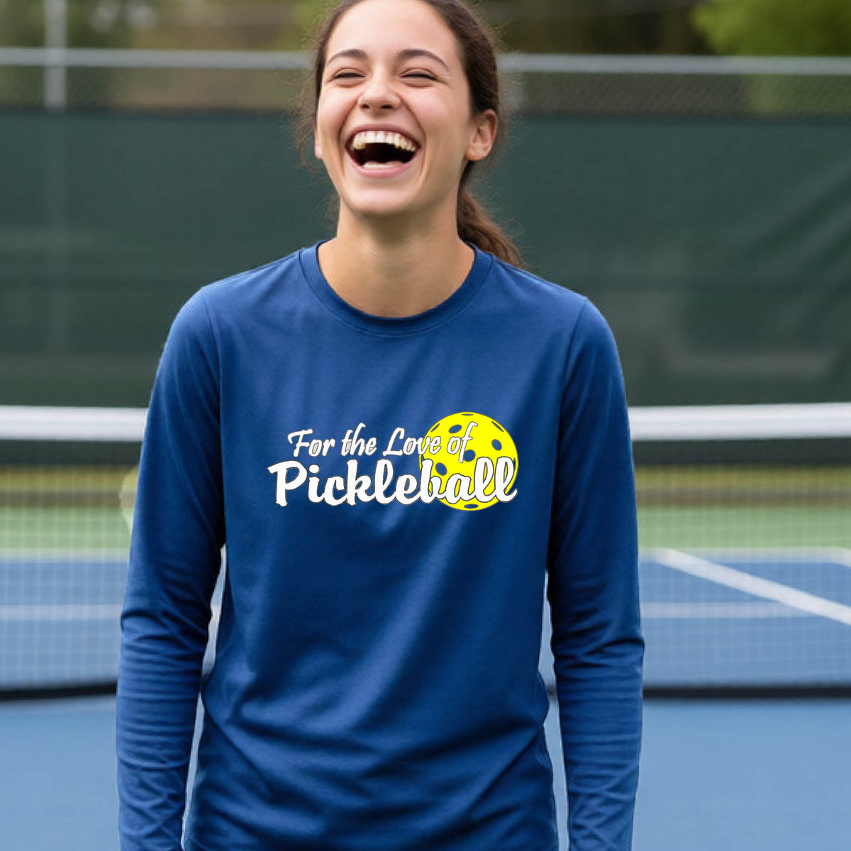 A closeup of a young person wearing a royal blue long-sleeve athletic shirt with a pickleball graphic on the left bottom left of ‘FOR THE LOVE OF PICKLEBLL’ in white with a yellow pickleball behind it.  