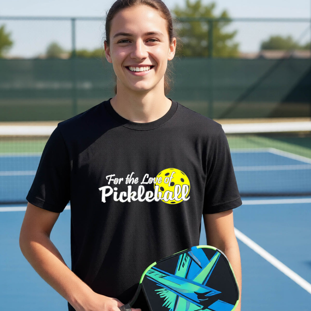 A closeup of a young person wearing a black short-sleeve athletic shirt with a pickleball graphic on the left bottom left of ‘FOR THE LOVE OF PICKLEBLL’ in white with a yellow pickleball behind it.  