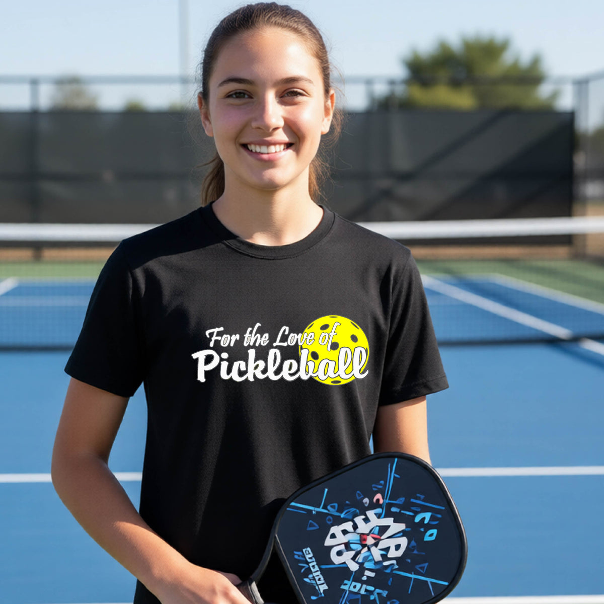 
A closeup of a young person wearing a black short-sleeve athletic shirt with a pickleball graphic on the left bottom left of ‘FOR THE LOVE OF PICKLEBLL’ in white with a yellow pickleball behind it.  
