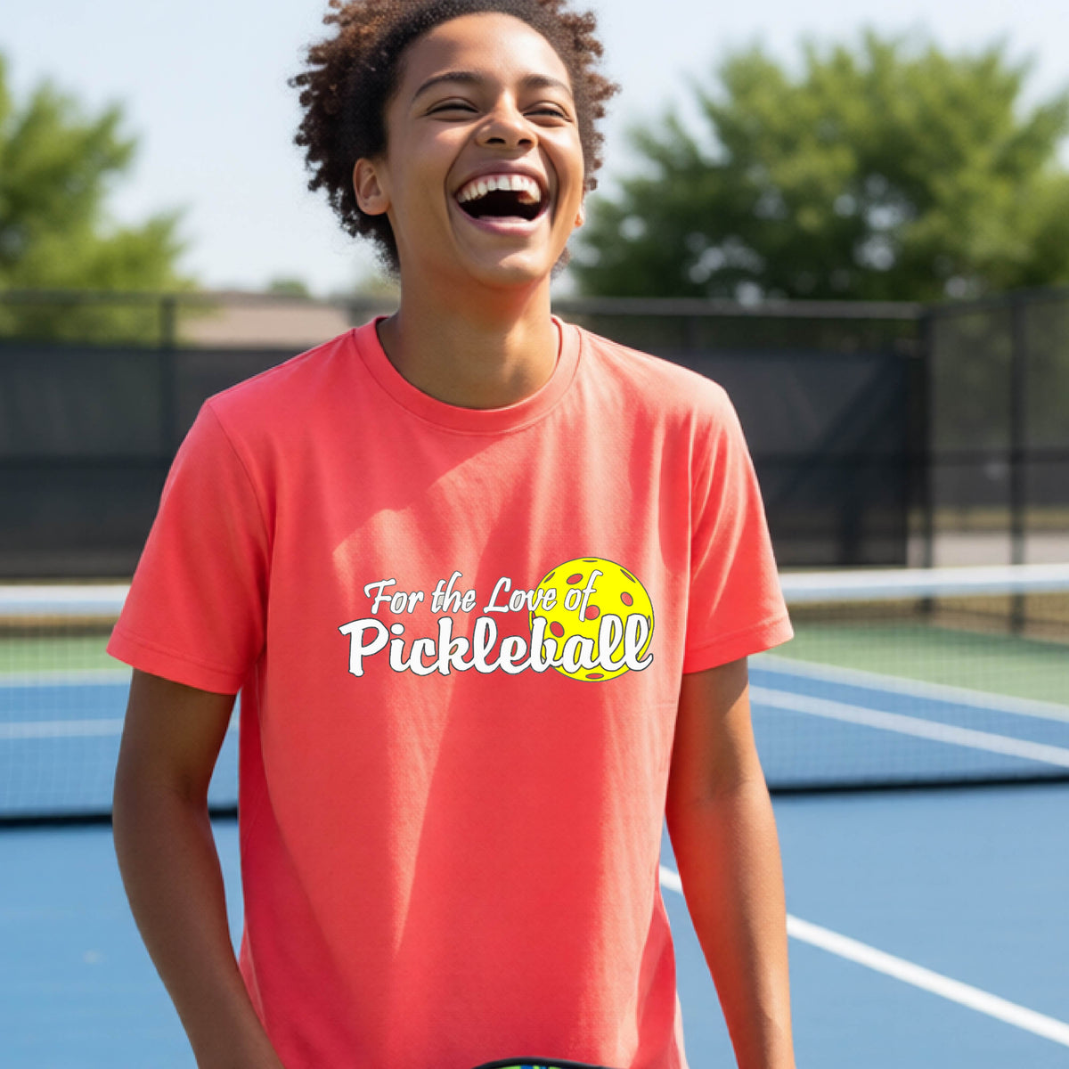 
A closeup of a young person wearing a coral pink short-sleeve athletic shirt with a pickleball graphic on the left bottom left of ‘FOR THE LOVE OF PICKLEBLL’ in white with a yellow pickleball behind it.  
