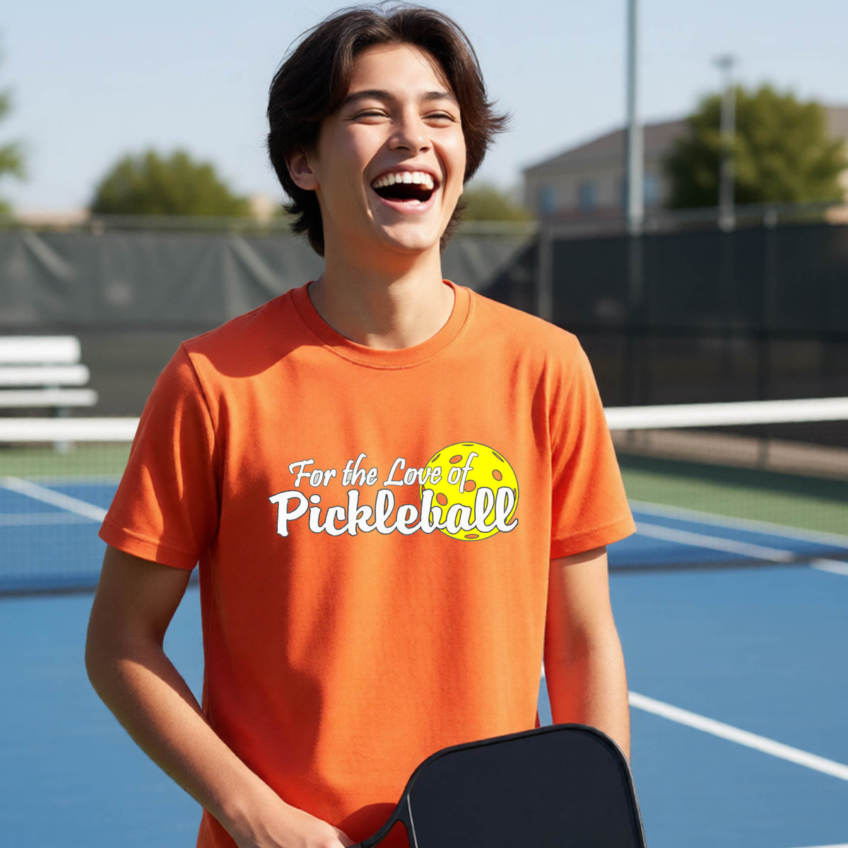 
A closeup of a young person wearing a orange short-sleeve athletic shirt with a pickleball graphic on the left bottom left of ‘FOR THE LOVE OF PICKLEBLL’ in white with a yellow pickleball behind it.  
