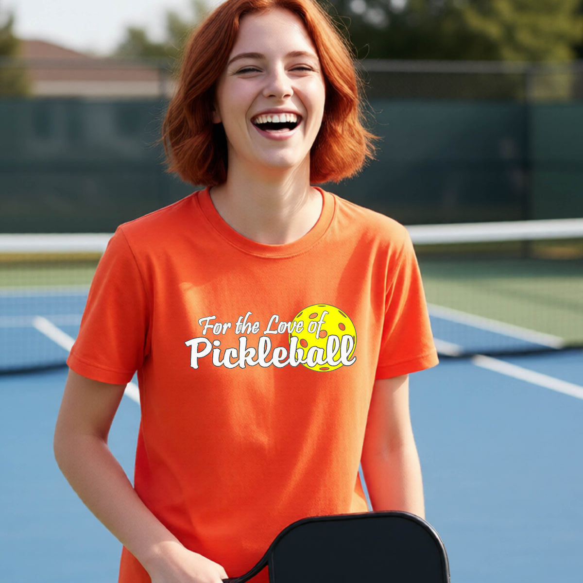 
A closeup of a young person wearing a orange short-sleeve athletic shirt with a pickleball graphic on the left bottom left of ‘FOR THE LOVE OF PICKLEBLL’ in white with a yellow pickleball behind it.  
