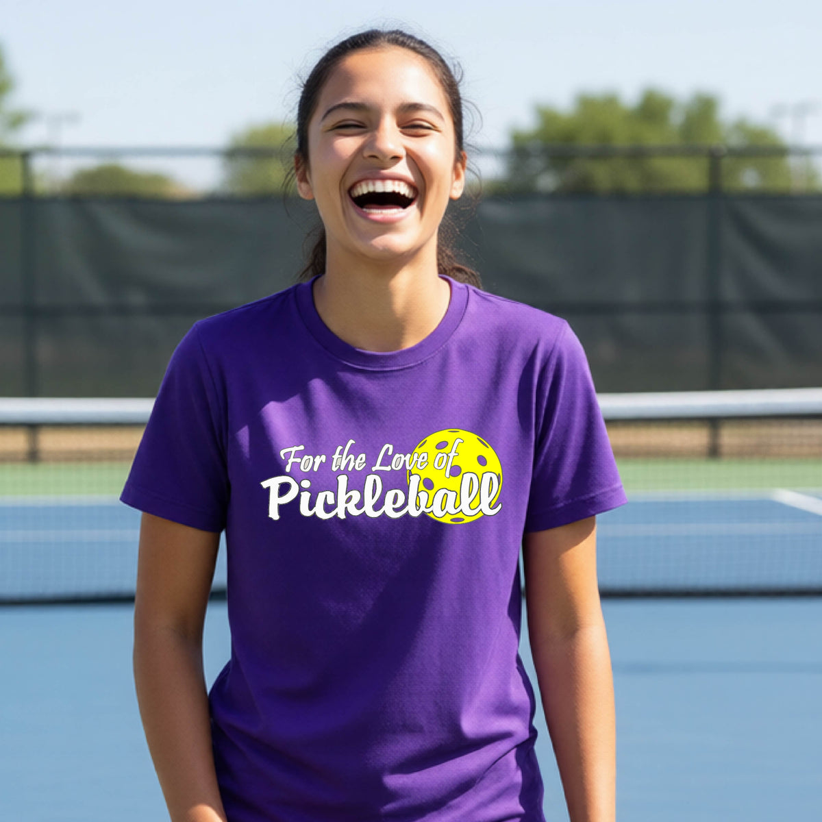 
A closeup of a young person wearing a purple short-sleeve athletic shirt with a pickleball graphic on the left bottom left of ‘FOR THE LOVE OF PICKLEBLL’ in white with a yellow pickleball behind it.  
