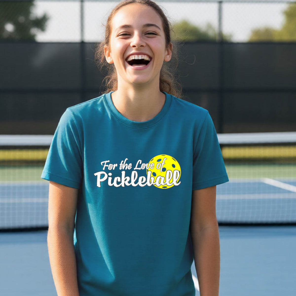 
A closeup of a young person wearing a teal short-sleeve athletic shirt with a pickleball graphic on the left bottom left of ‘FOR THE LOVE OF PICKLEBLL’ in white with a yellow pickleball behind it.  
