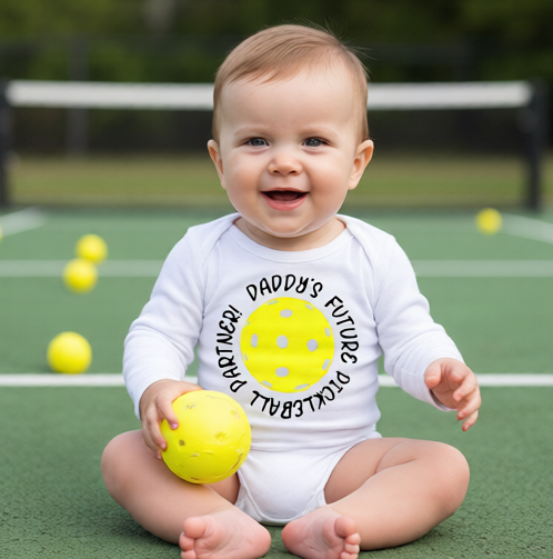 A baby sitting on a pickleball court wearing a white bodysuit with a yellow pickleball graphic and the text 'DADDY'S FUTURE PICKLEBALL PARTNER'. 