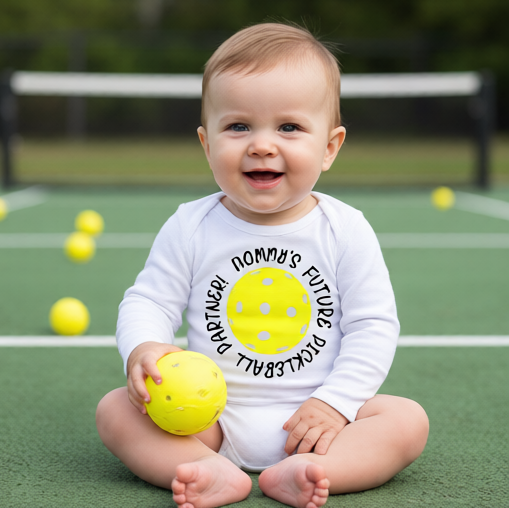 A baby sitting on a pickleball court wearing a white bodysuit with a yellow pickleball graphic and the text 'MOMMY'S FUTURE PICKLEBALL PARTNER'. 