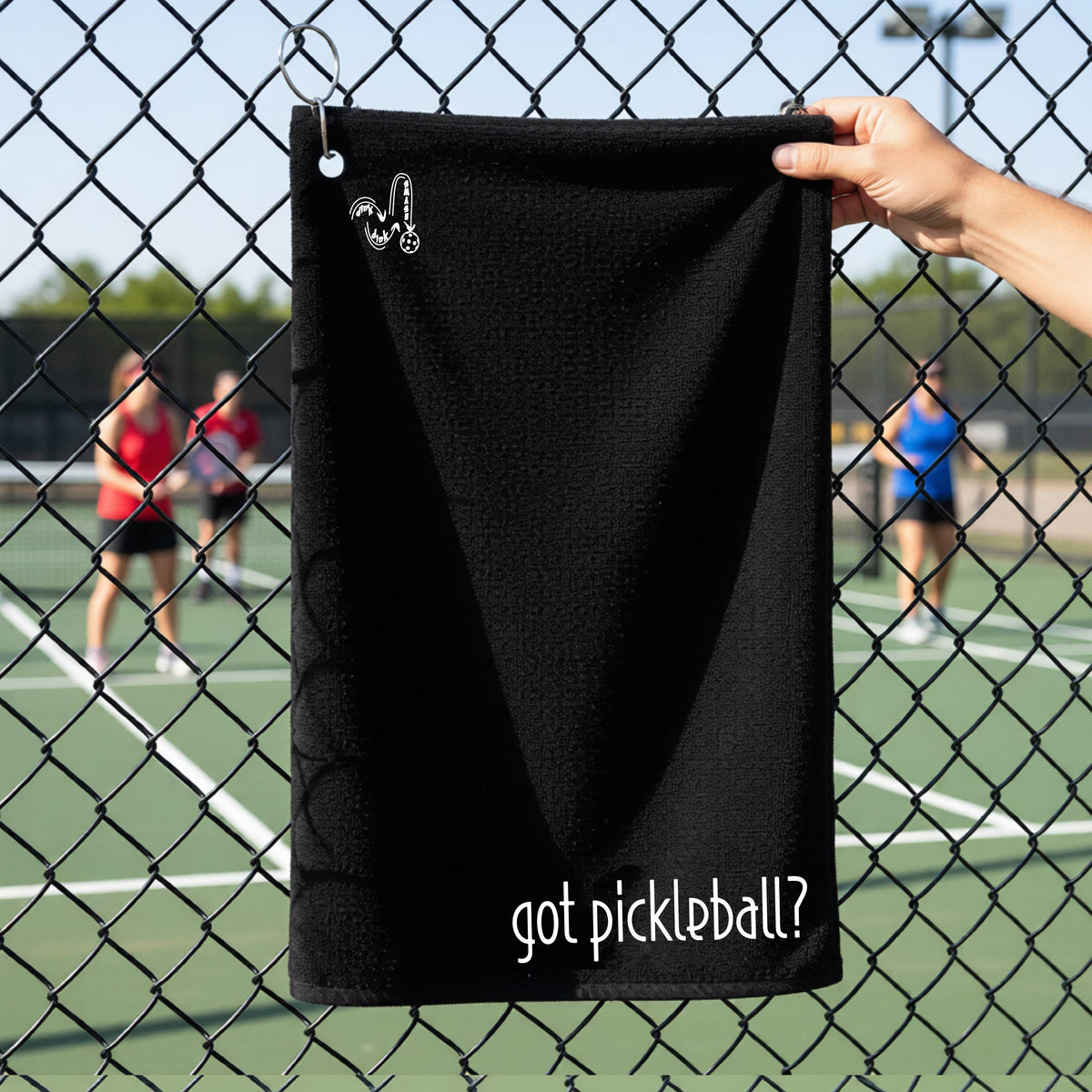 A close-up of a black terry cloth court towel hanging on a pickleball court fence with a white logo of ‘DINK DINK SMASH’ in the top left corner, and a white text of ‘got pickleball’ in the bottom right of the towel. 
