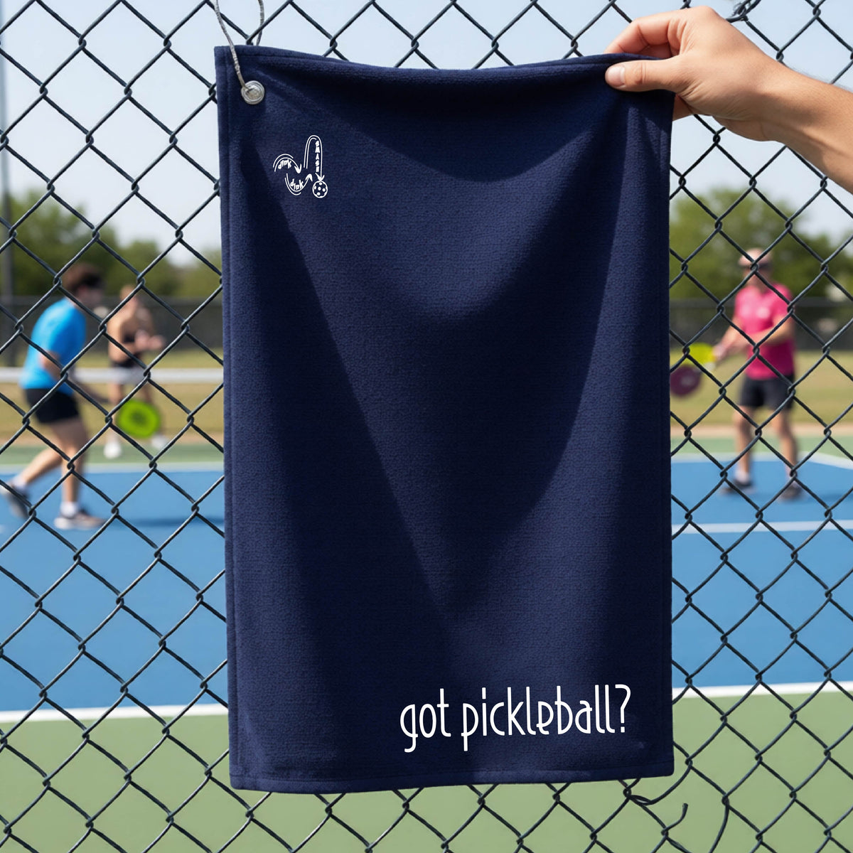 A close-up of a navy blue terry cloth court towel hanging on a pickleball court fence with a white logo of ‘DINK DINK SMASH’ in the top left corner, and a white text of ‘got pickleball’ in the bottom right of the towel. 