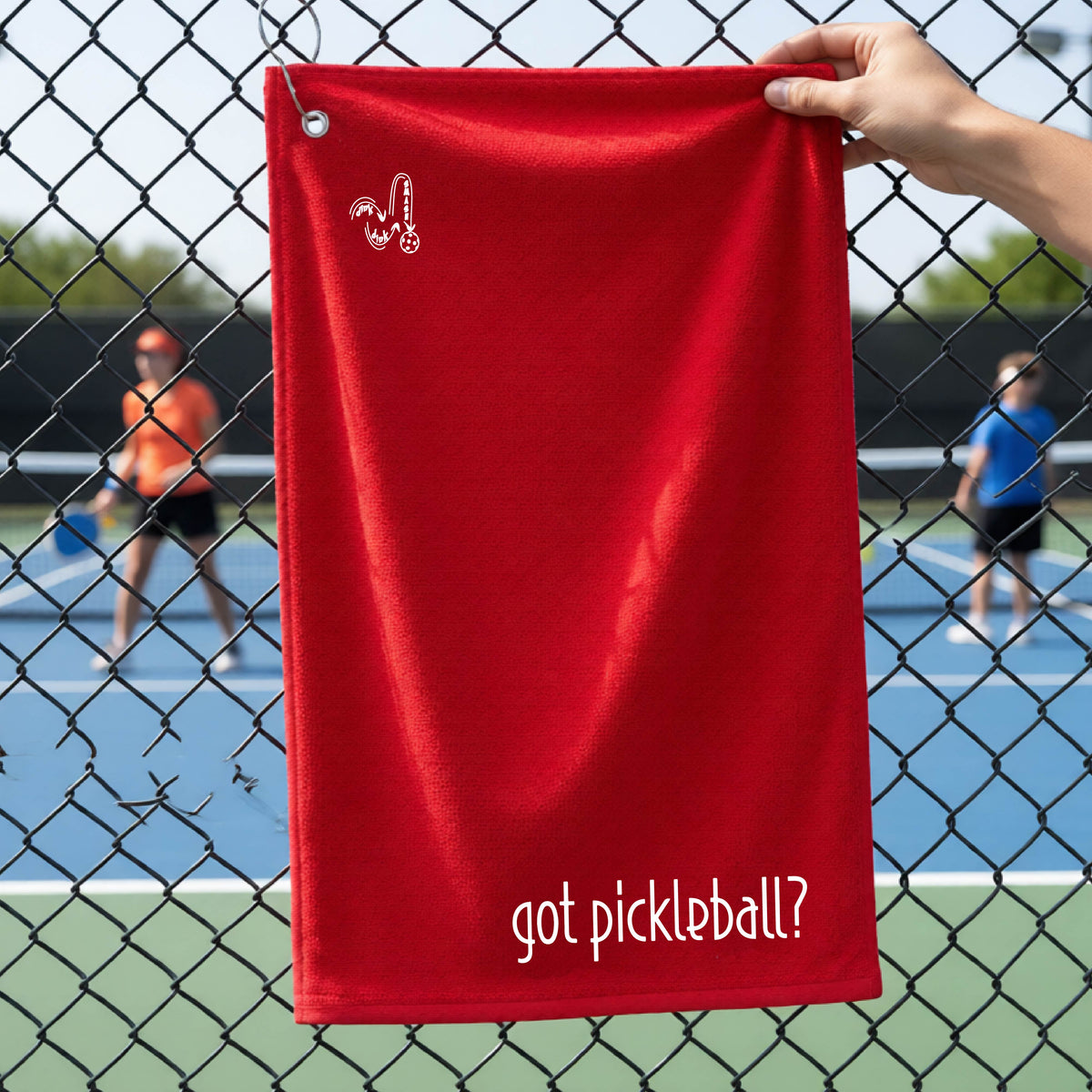 A close-up of a red terry cloth court towel hanging on a pickleball court fence with a white logo of ‘DINK DINK SMASH’ in the top left corner, and a white text of ‘got pickleball’ in the bottom right of the towel. 