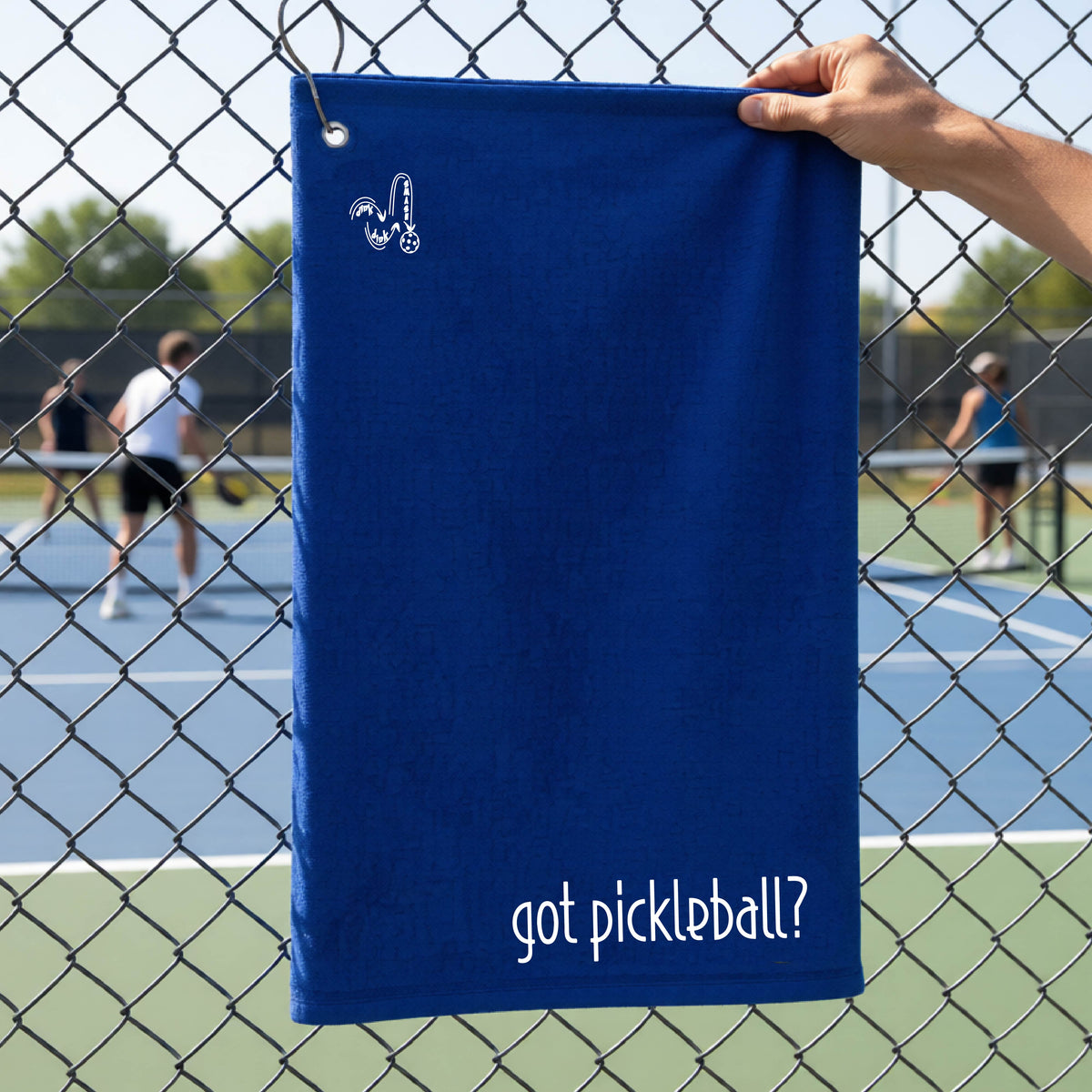 A close-up of a royal blue terry cloth court towel hanging on a pickleball court fence with a white logo of ‘DINK DINK SMASH’ in the top left corner, and a white text of ‘got pickleball’ in the bottom right of the towel. 