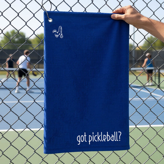 A close-up of a royal blue terry cloth court towel hanging on a pickleball court fence with a white logo of ‘DINK DINK SMASH’ in the top left corner, and a white text of ‘got pickleball’ in the bottom right of the towel. 