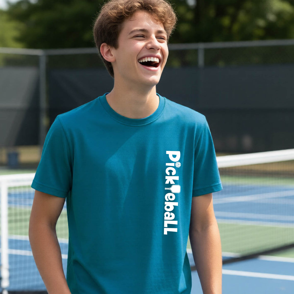 A young person on a pickleball court seen from behind, wearing a teal t-shirt with 'Pickleball' printed vertically in white on the front.