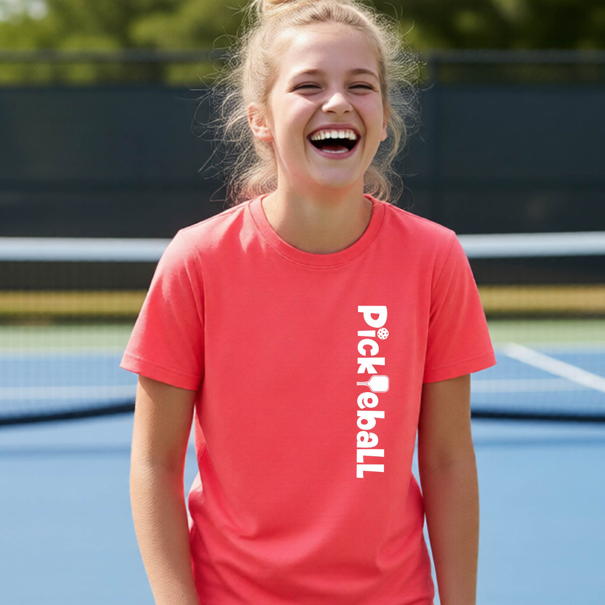 A young person on a pickleball court seen from behind, wearing a coral pink t-shirt with 'Pickleball' printed vertically in white on the front.