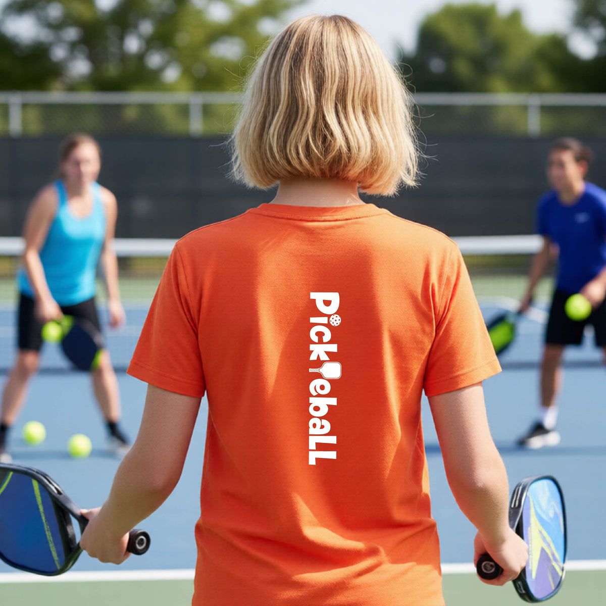 A young person on a pickleball court seen from behind, wearing an orange t-shirt with 'Pickleball' printed vertically in white on the back.